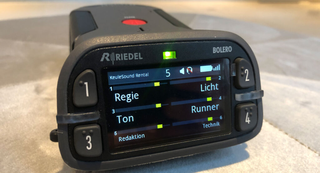 Eine Nahaufnahme eines Riedel Bolero Handfunkgerätes für intercom, oben sieht man den Roten Punkt eine Taste, Auf dem Display kann man die verschiedenen Intercomkreise lesen, jeder Kreis hat einen Button welcher die Zahl 1 , 2 , 3, 4 trägt. Die Grüne LED in der Mitte signalisiert das das Gerät bereit ist. Intercom Riedel Mieten, Hier das Riedel Bolero Beltpack