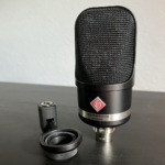 Ein Mikrofon stehend auf dem XLR Anschluss, das Mikrofon ist schwarz mit einem großen roten Logo auf dem Neumann steht. Daneben liegt eine Mikrofonhalterung. Neumann TLM 107 Multipattern Großmembranmikrofon