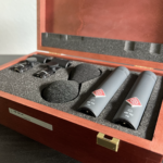 Eine braune Holzkiste in welcher Mikrofone liegen, daneben liegen auch zwei Windschutz und zwei Stativklemmen. Stereo Set Neumann KM 184