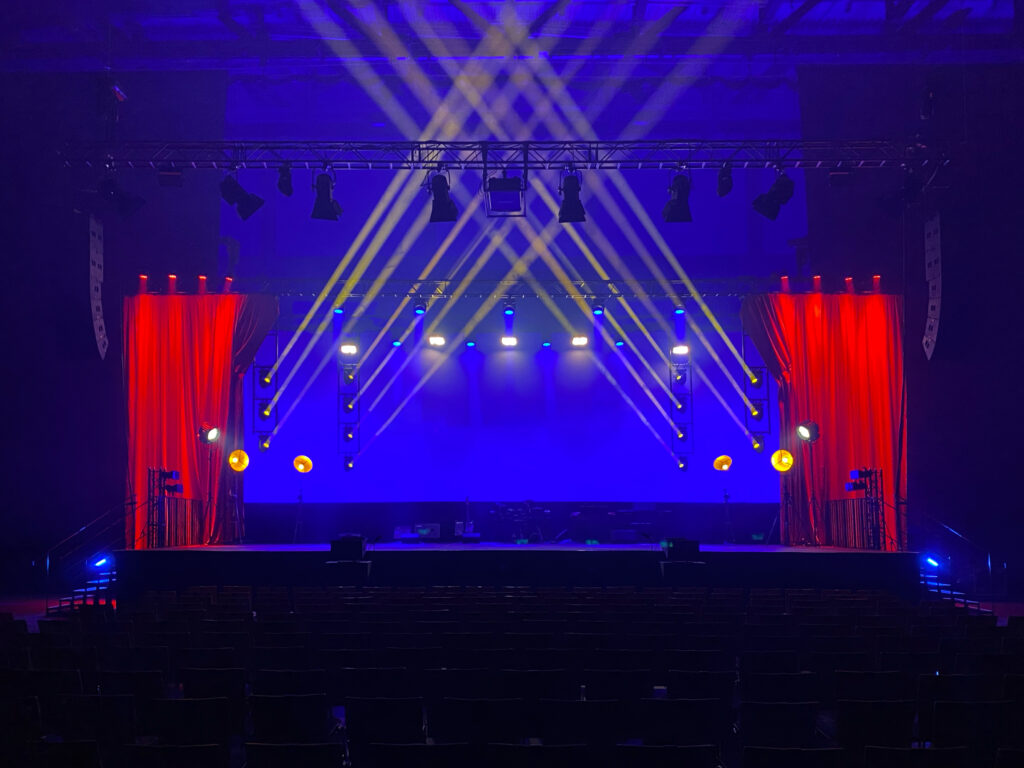 Eine Bühne Frontal aufgenommen der Hintergrund ist blau beleuchtet rechts und links Gelbe Lichtkegel die nach Oben in die Mitte zeigen. Rechts und Links der Bühne ist mit rotem Licht ausgeleuchtet. Gala Veranstaltung Riedel Bolero @ Wetzlar Intercom 10 er Set mieten