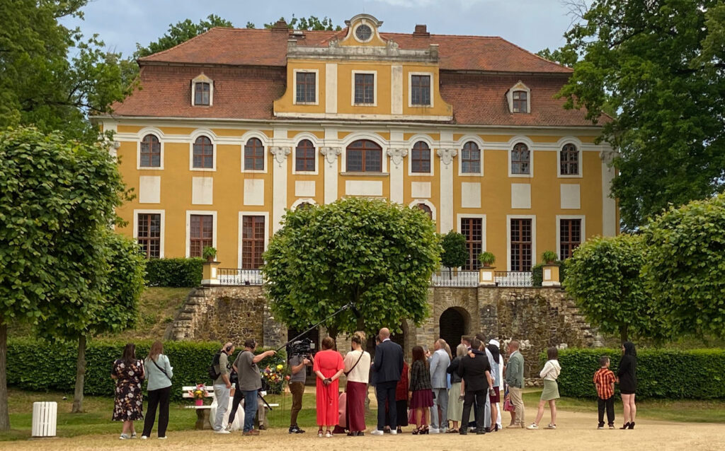 Im Hintergrund ein Schloss, vor dem Schloss ein Baum vor dem eine Hochzeitsgesellschaft steht. Intercom bei "Hochzeit auf den ersten Blick"