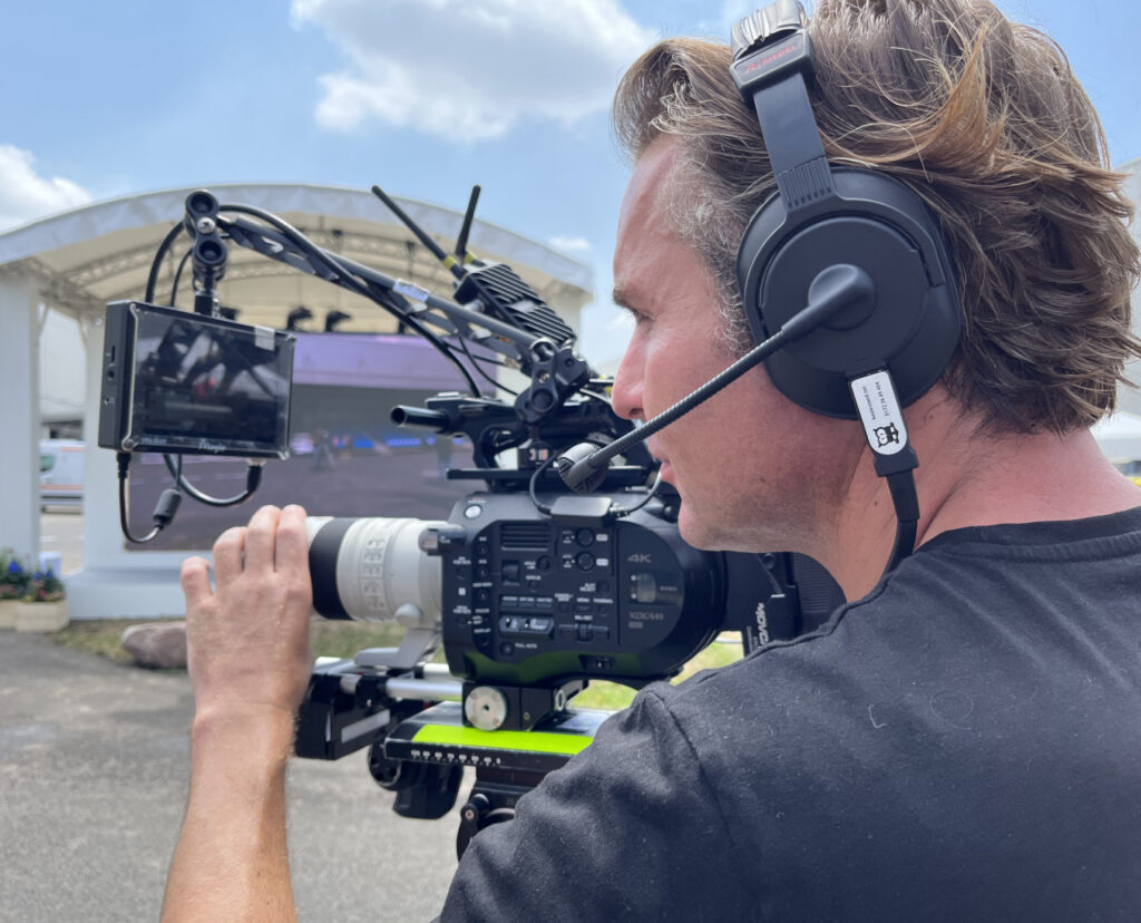 Ein Mann mit einem Headset auf dem Kopf und einer Kamera in der Hand. Im Hintergrund eine Bühne. Riedel Bolero Intercom bei der Präsentation des I4 in Leipzig