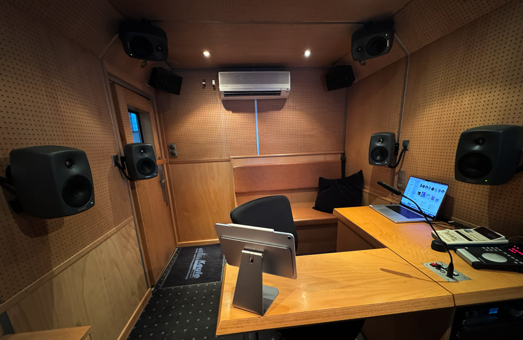 Ton-Ü-Wagen von innen, viel Holz mit Löchern für eine akustische Optimierung. Mehrere graue Lautsprecher an Wand und Decke für Abmischung in Dolby Atmos